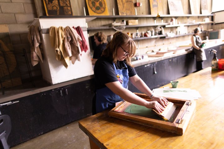 A student is screen printing on a large wooden table in an art studio, concentrating on the process. Shelves filled with art supplies and canvases line the wall behind her, while other students work in the background.